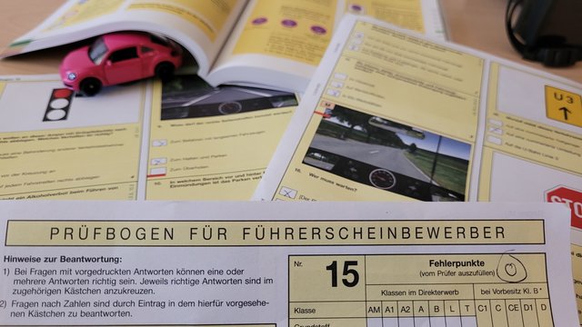 Ein Ausschnitt von einem Prüfbogen und Lehrbüchern für die Führerscheinprüfung. Unter einem Buch sieht man ein kleines rotes Spielzeugauto.
