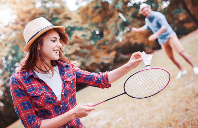 Eine junge Frau mit Hut macht einen Badminton-Aufschlag, im Hintergrund steht ein junger Mann mit Schläger und wartet auf den Rückschlag.