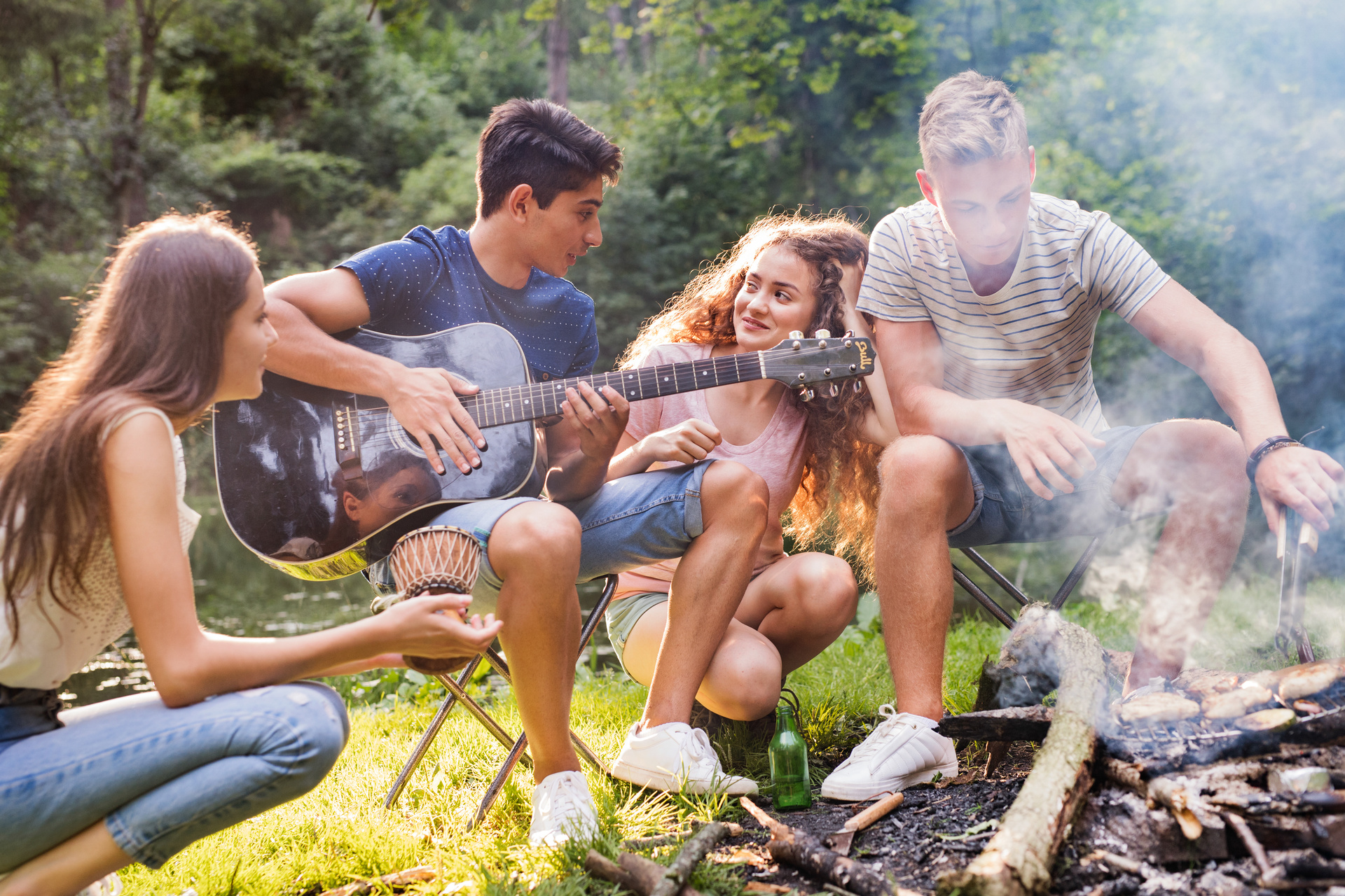 Zwei junge Frauen und zwei junge Männer sitzen gemeinsam an einem Lagerfeuer bei Tageslicht – einer von ihnen spielt Gitarre.