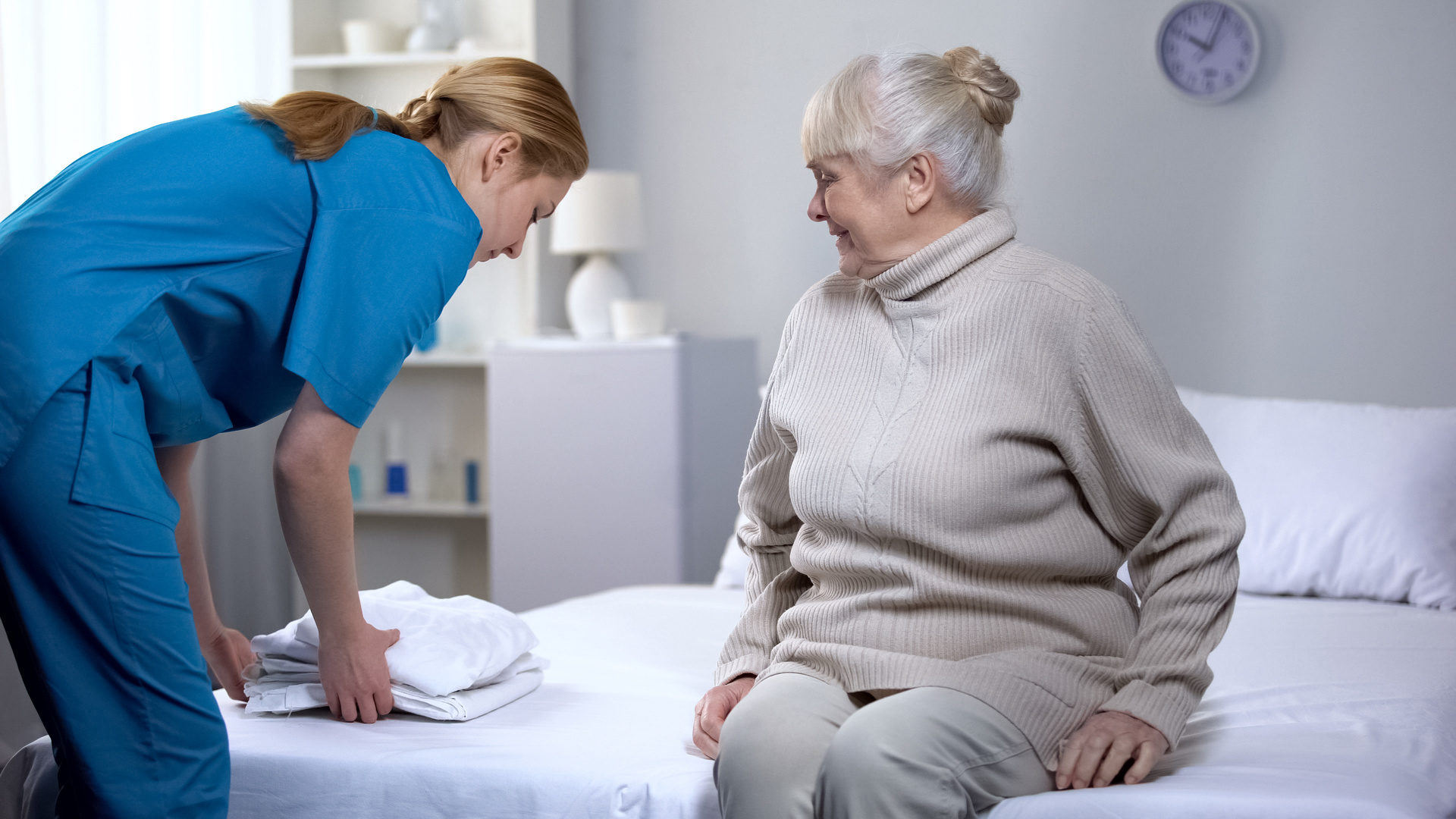 Eine Frau in blauer Arbeitskleidung legt frische Wäsche auf ein Bett, auf dem eine ältere Dame sitzt und sie freundlich anschaut.