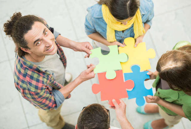 Vier Personen halten jeweils ein passendes Puzzleteil in der Mitte zusammen. Eine Person blickt nach oben in die Kamera und lächelt.