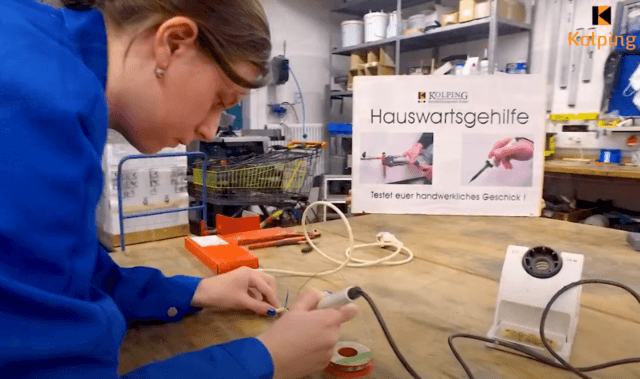 Eine Teilnehmerin im Blaumann steht an einem Werktisch und lötet konzentriert ein Bauteil.