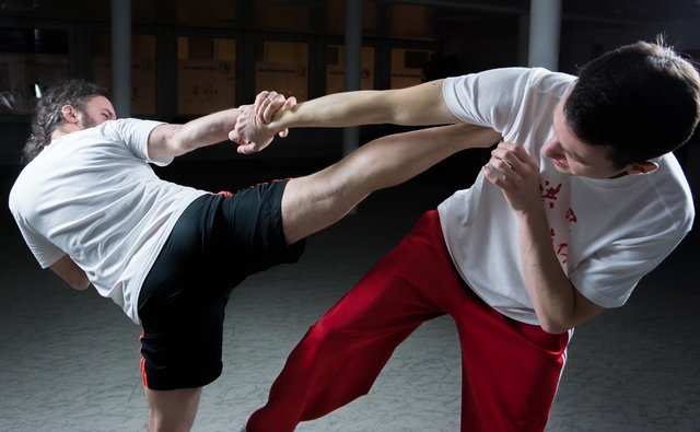 Zwei Männer beim Kampfsport: Während sie sich an den Händen greifen, hat einer sein Bein bereits in der Luft und führt einen Tritt aus